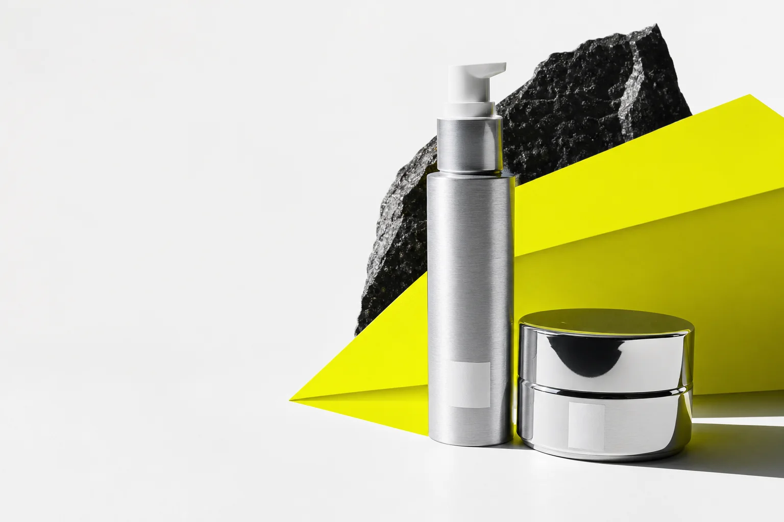 Silver MATERIA serum bottle and cream jar arranged with acid-yellow paper and black mineral stone